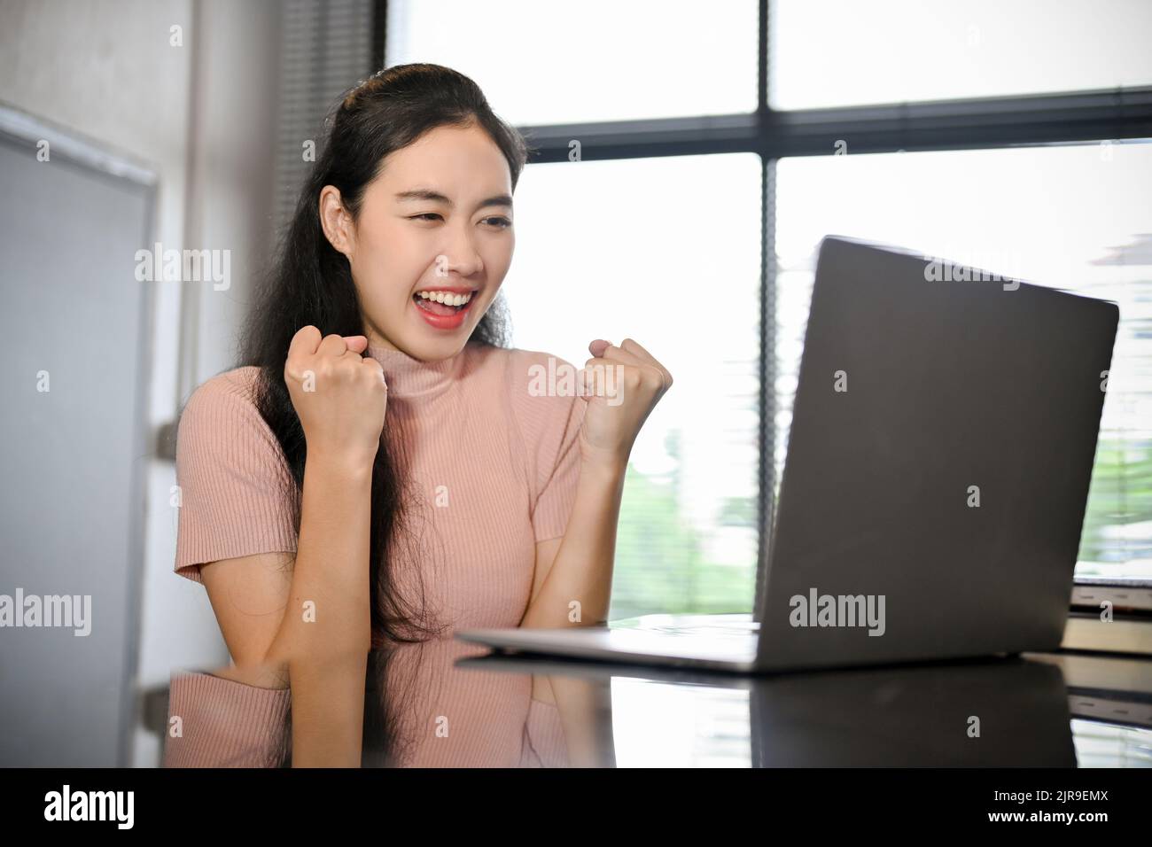 Excited young Asian female looking at laptop screen, happy after receiving a job offer email, a work promotion, or passing the hardest school test. Stock Photo