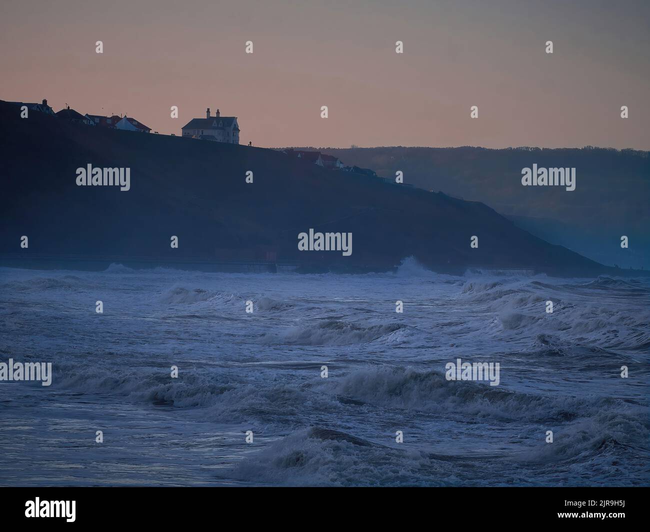 Angry storm waves crash into the bay between Whitby and Sandsend on the North Sea coast, as the winter sun sets behind the headland and cliffs. Stock Photo