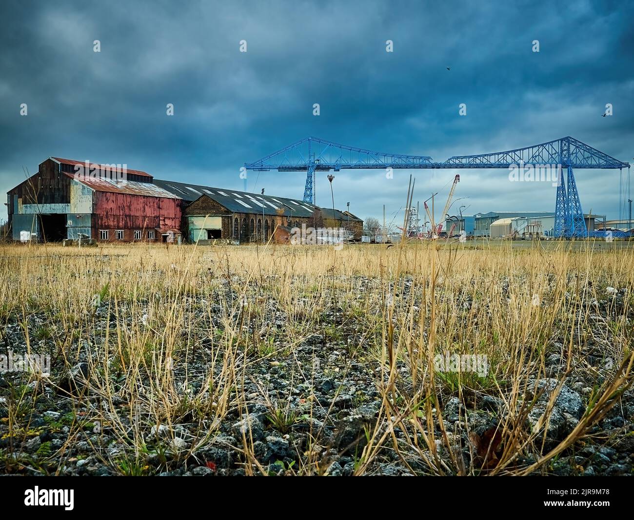 Low angle view of a weed-strewn waste, connected to derelict, corroded warehouses with a threatening sky and the emblematic Transporter Bridge behind. Stock Photo