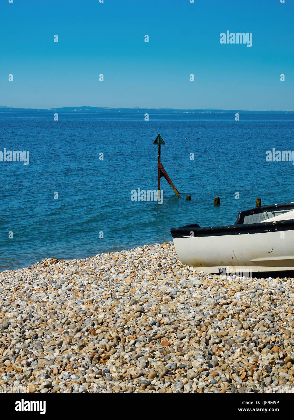 A small boat pulled onto a deserted shingle beach near a marker buoy and a rippled blue sea, under a clear summer sky. Stock Photo