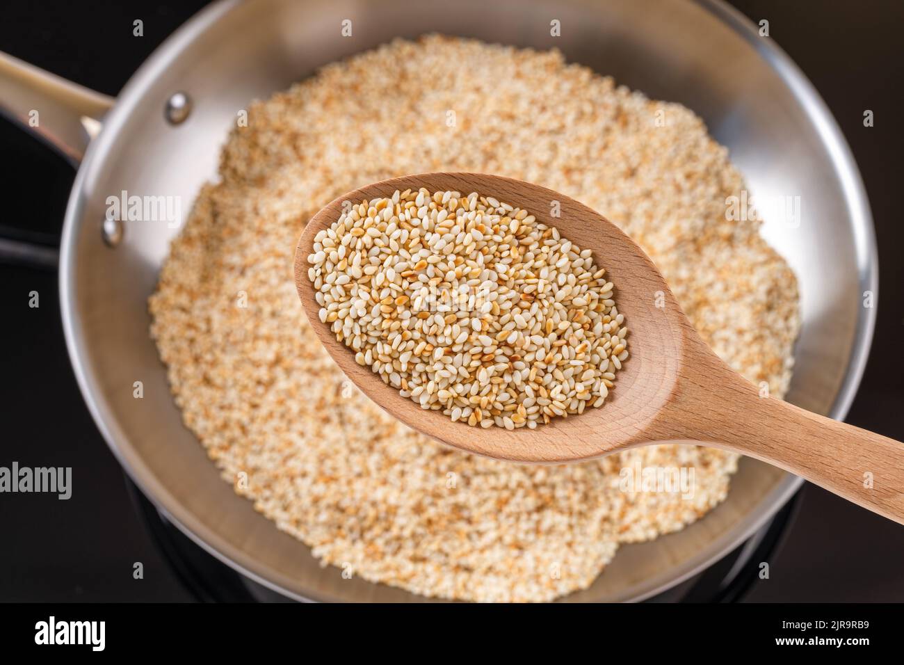 Toasted sesame seeds in a wooden spoon over stainless steel skillet. Roasting organic benne grains in a frying pan. Sesamum indicum as calcium source. Stock Photo