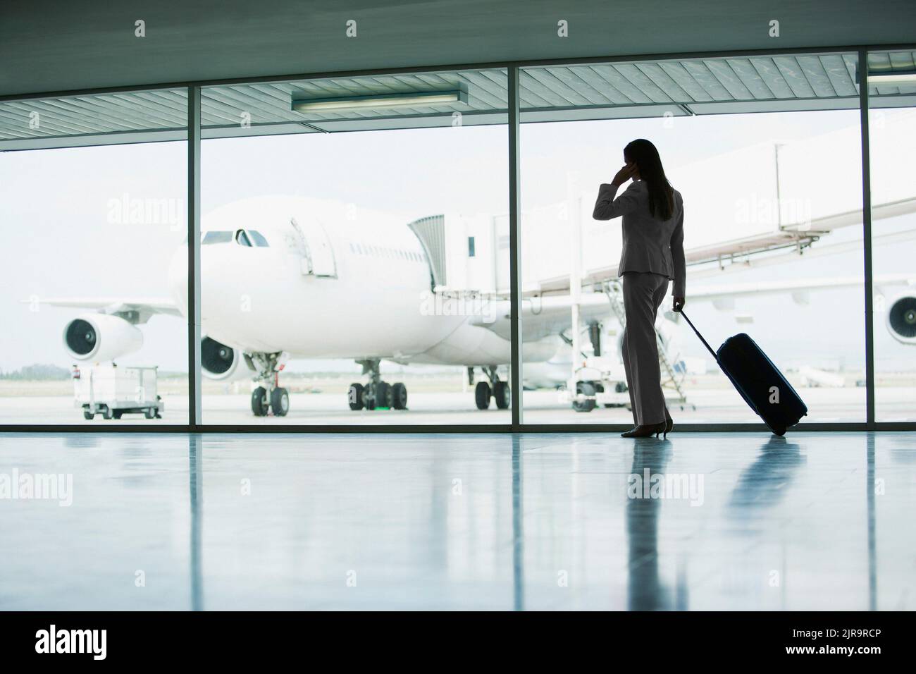 Businesswoman with a wheeled suitcase waiting for her flight and making a phone call with her smartphone. Runway and plane on the tarmac viewed from t Stock Photo