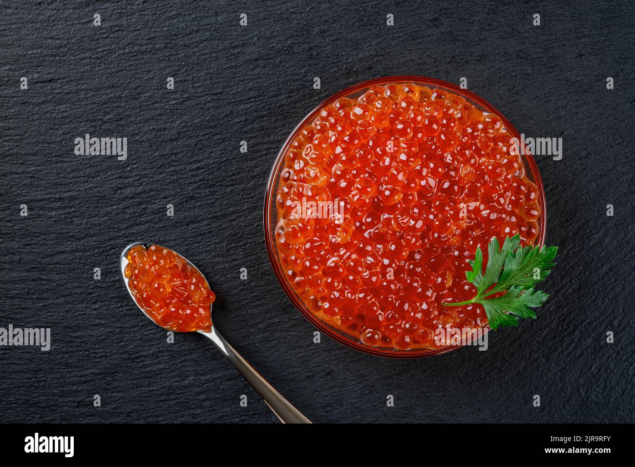 Delicious salmon caviar in a glass bowl over black slate background. Close-up of bowl and spoon full of trout red caviar. Salted salmon roe. Top view. Stock Photo