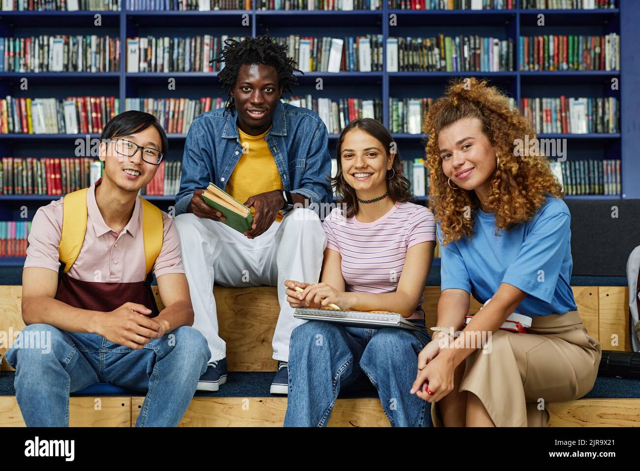 Front view at diverse group of students in library smiling happily at camera against bookshelves Stock Photo