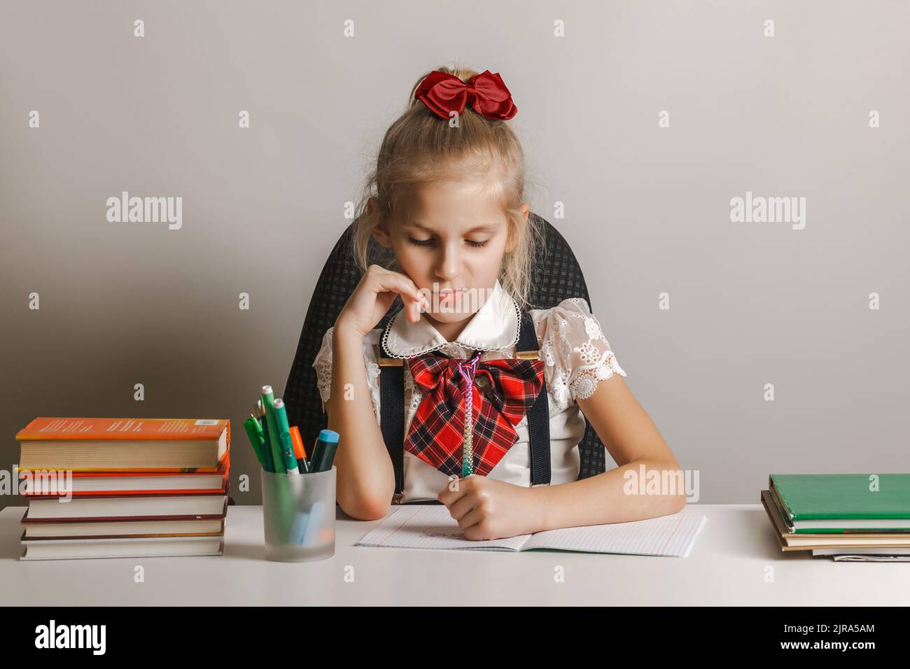 A little girl with a dissatisfied face in a school uniform draws in a notebook with her left hand, she does not like what she is doing. Stock Photo