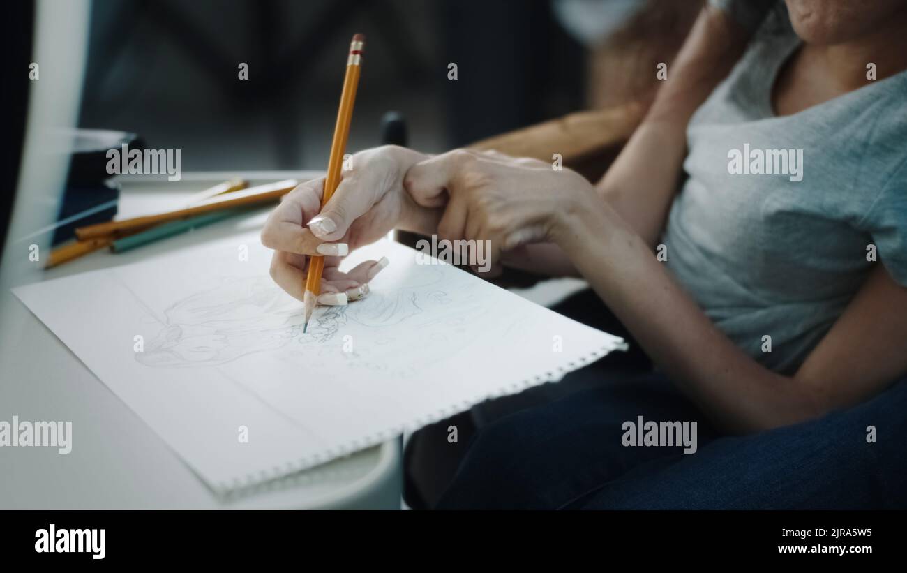 Woman with disability in a wheelchair sitting at the desk and drawing a sketch, with pencil on a sheet of paper and records it on video or makes a live stream on the social network Stock Photo