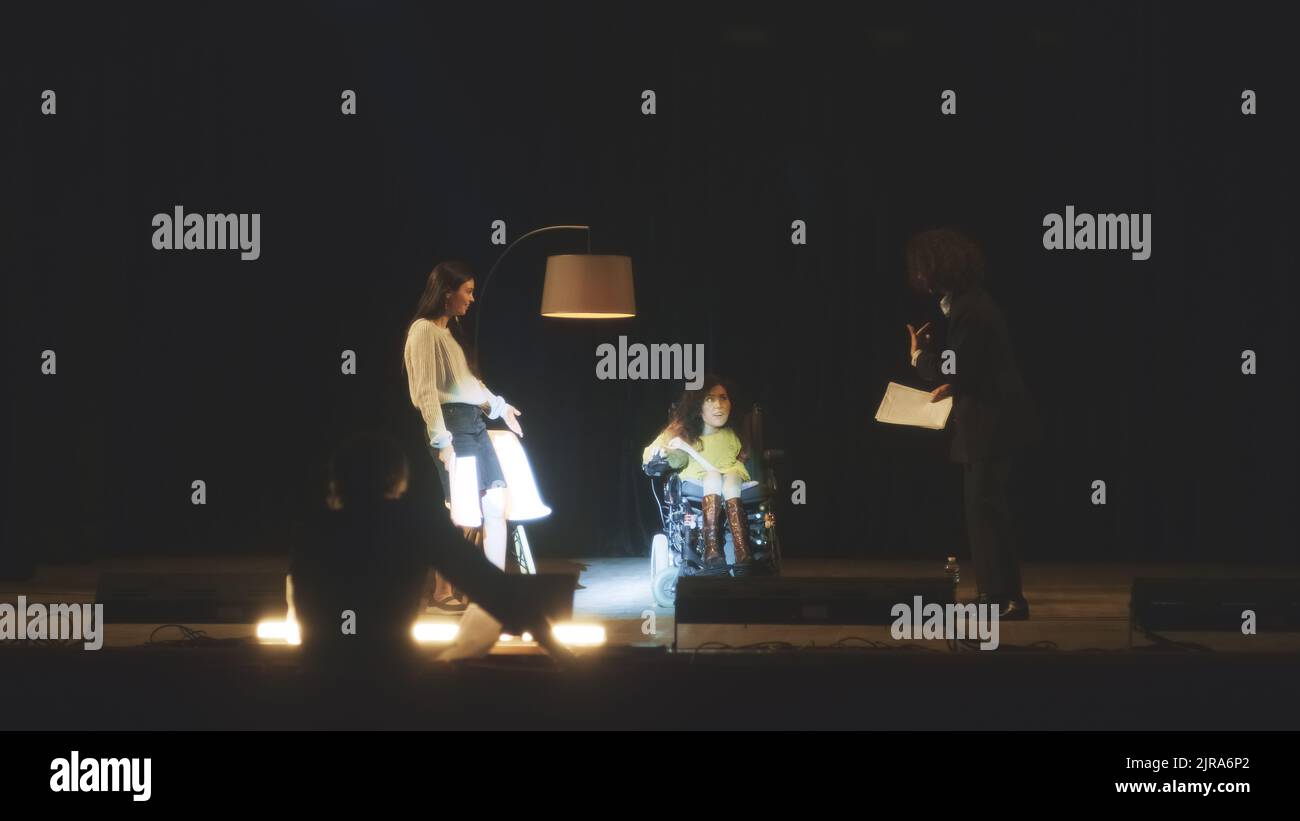 A troupe of actors with disabled woman in a wheelchair rehearsing a comedy play on a stage in a dark theater with a director together Stock Photo