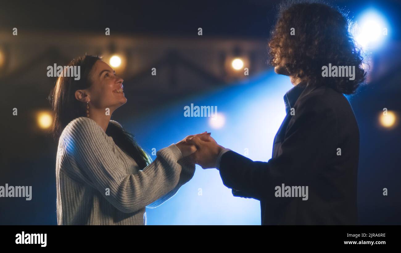 Male and female theater actors holding hands during a rehearsal of a romantic play on a stage illuminated by a spotlight Stock Photo