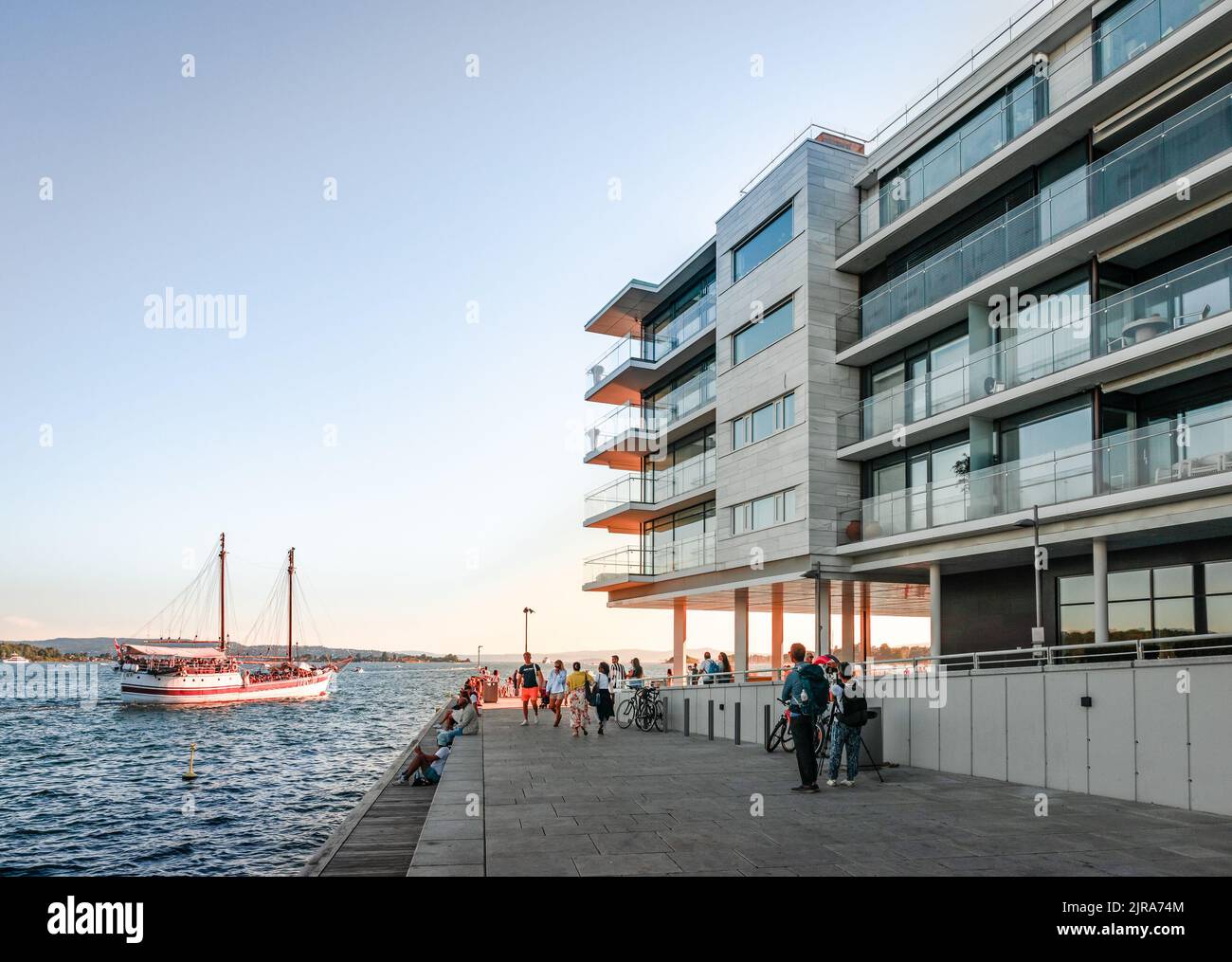 The waterfront of Tjuvholmen, Olso, Norway at sunset, with modern residential buildings facing the sea and a cruising boat in the background. Stock Photo