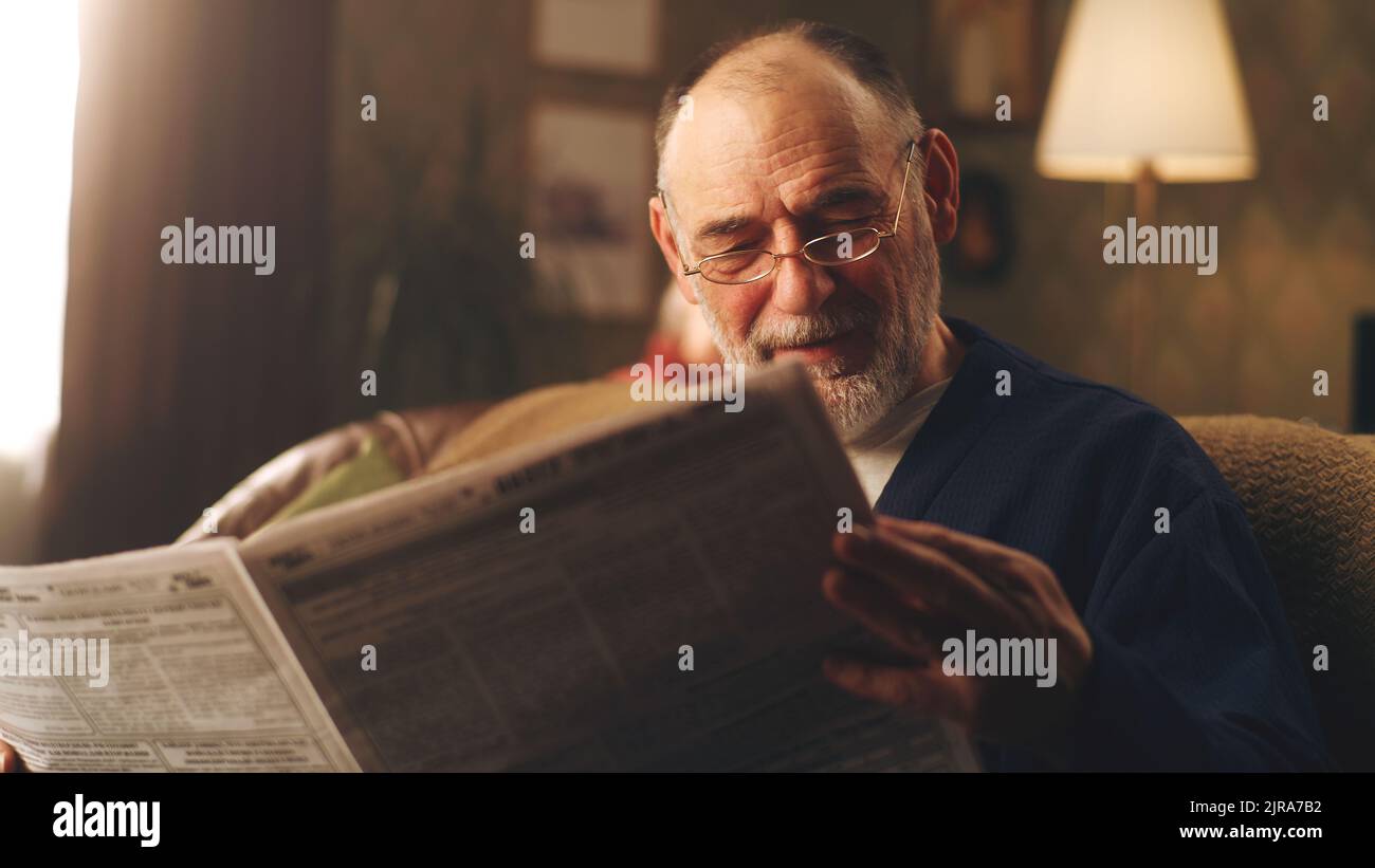 Cheerful senior man sitting on the couch in living room and laughing while watching latest news in the newspaper and reading jokes Stock Photo