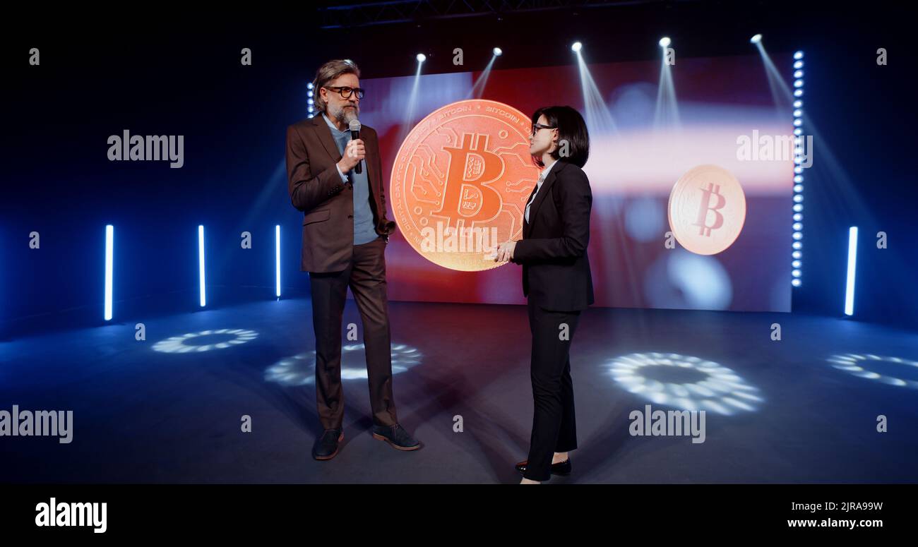 A female presenter in a suit interviewing a cryptocurrency coach on stage  in front of an