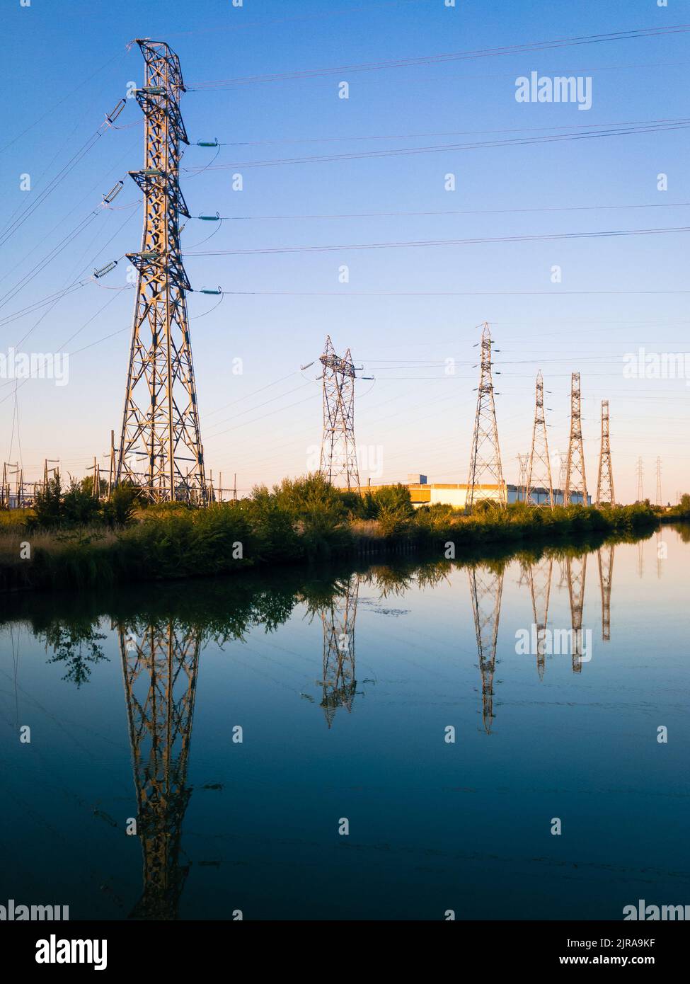 A row of metallic transmission towers of various shapes reflecting in the still water of a canal against blue sky at dusk. Stock Photo