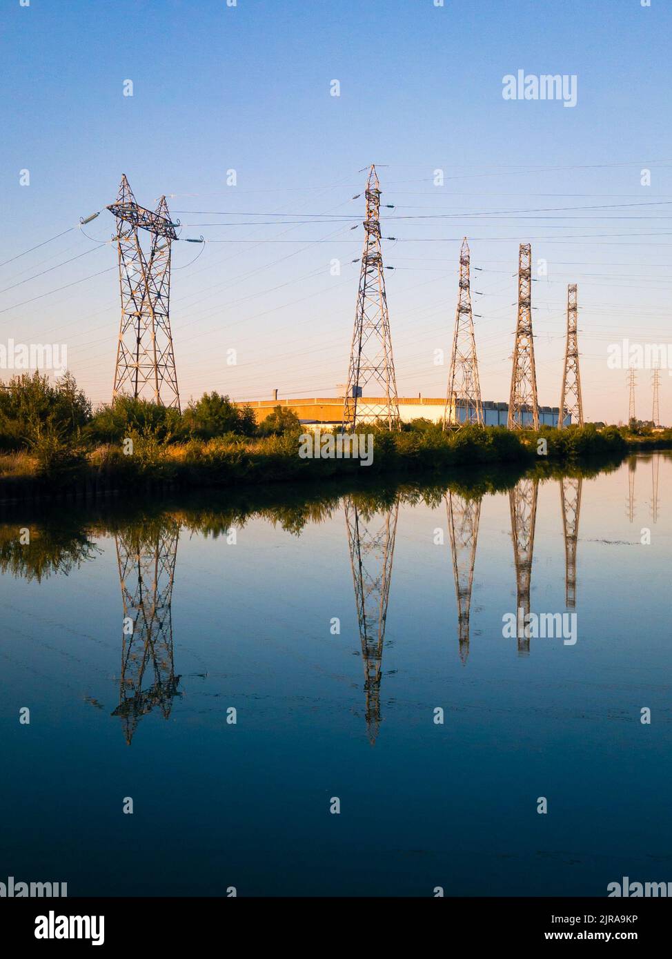 A row of metallic transmission towers of various shapes reflecting in the still water of a canal against blue sky at dusk. Stock Photo