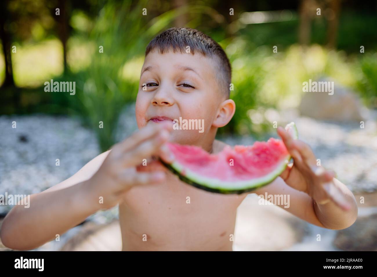 Little boy sitting near lake and eating watermelon on hot sunny day during summer vacation. Stock Photo