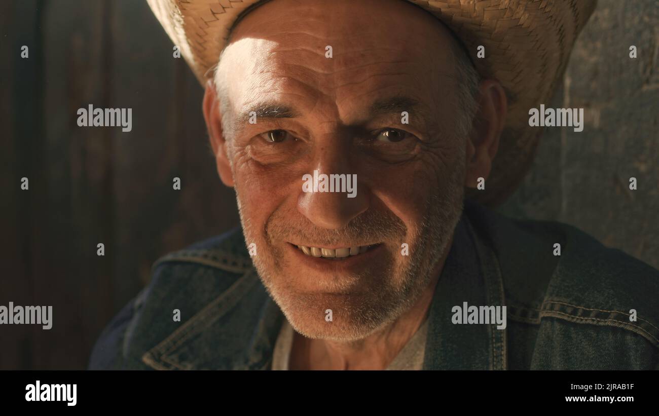 Glad senior man in straw hat smiling and looking at camera while working in sunlit barn on farm Stock Photo