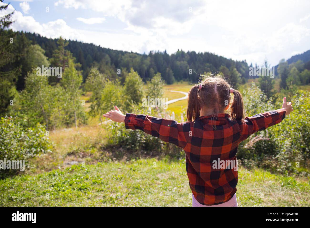 Caucasian child girl, back view, with open arms in a natural mountain landscape on a sunny day. Pian di Gembro natural park, Valtellina, Italy. Stock Photo