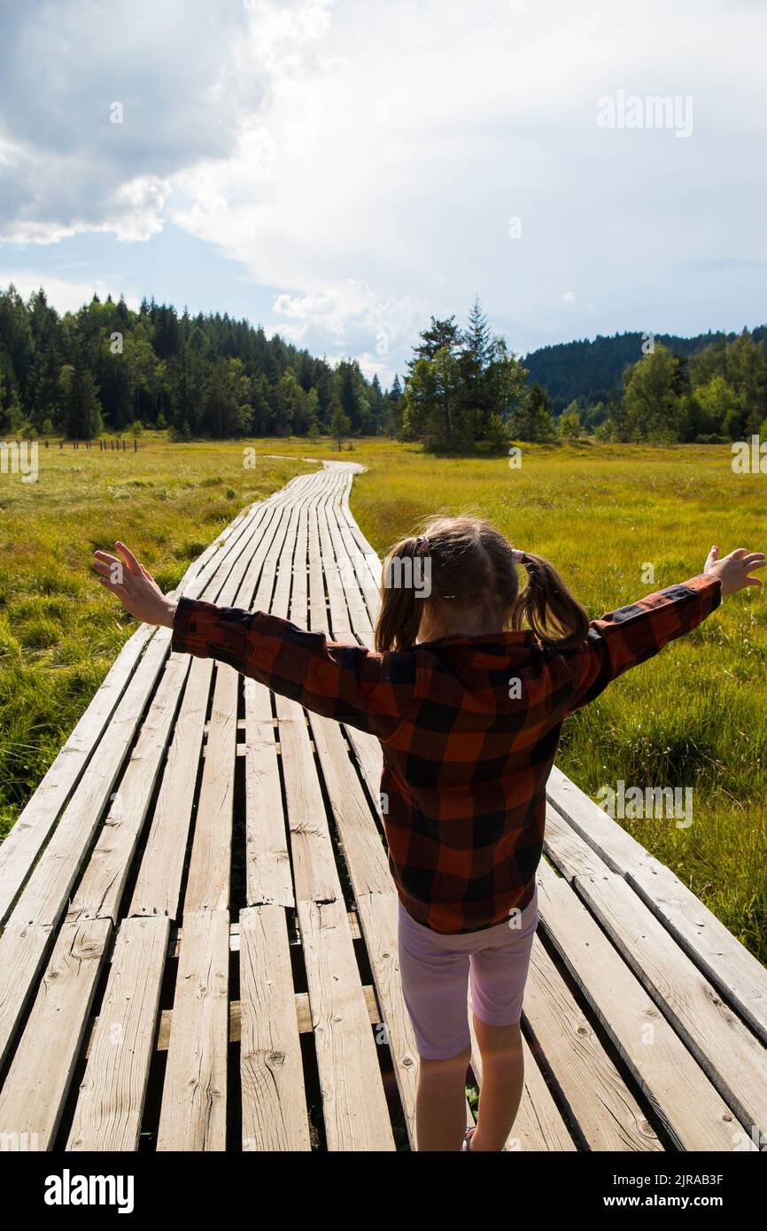 Caucasian child girl, back view, with open arms in a natural mountain landscape on a wooden path. Pian di Gembro natural park, Valtellina, Italy. Stock Photo