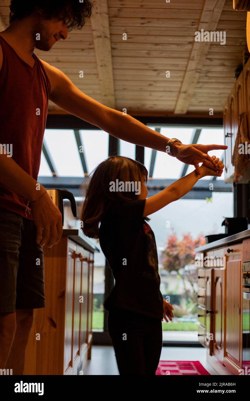 Caucasian young father and child girl dancing together in wooden kitchen. Happy simple moment, vertical shot. Stock Photo
