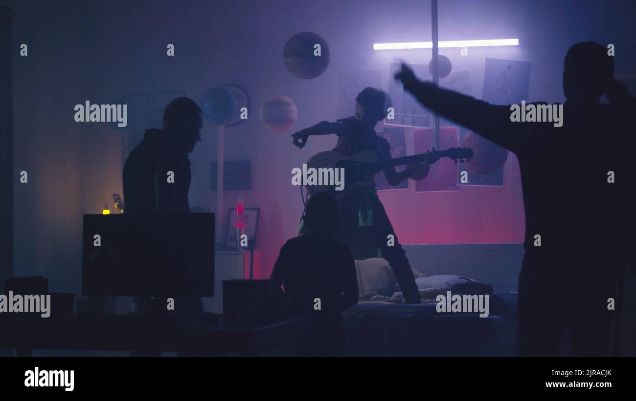Father and siblings dancing and applauding to teenager playing electric guitar under neon illumination during rock concert at night at home Stock Photo
