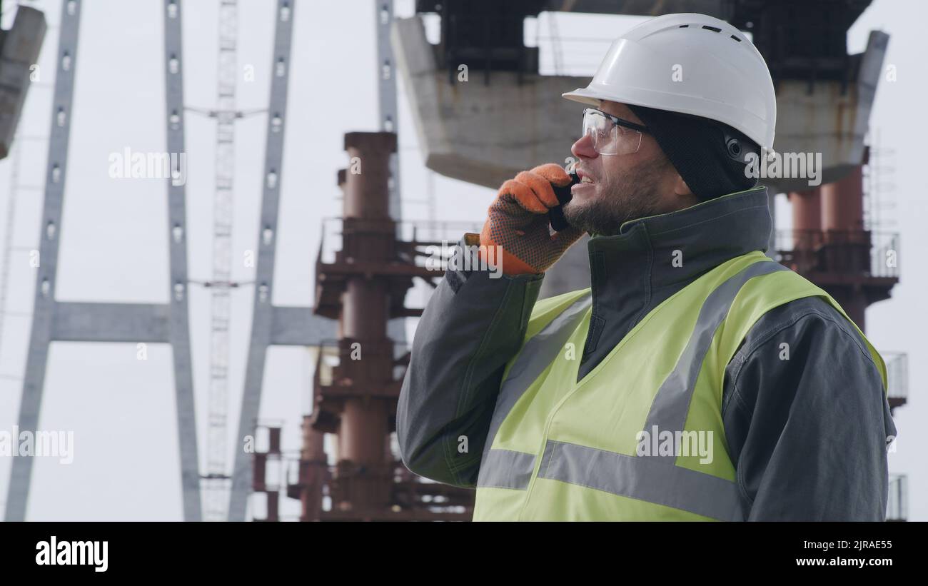 Man in hardhat and goggles inspecting unfinished bridge and talking on smartphone with boss during work on construction site Stock Photo