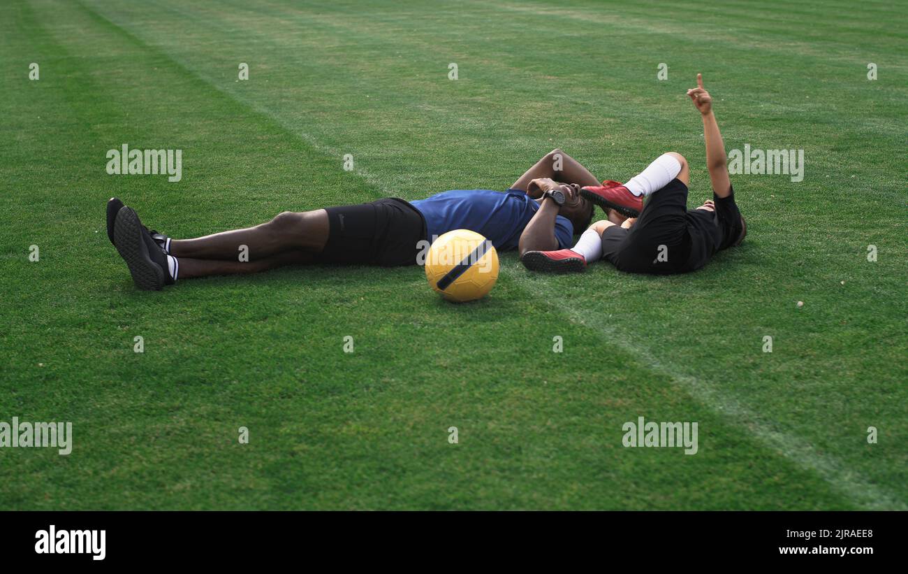 Pan around view of black man tickling mixed race boy and pointing at sky while lying on football field during break in training Stock Photo