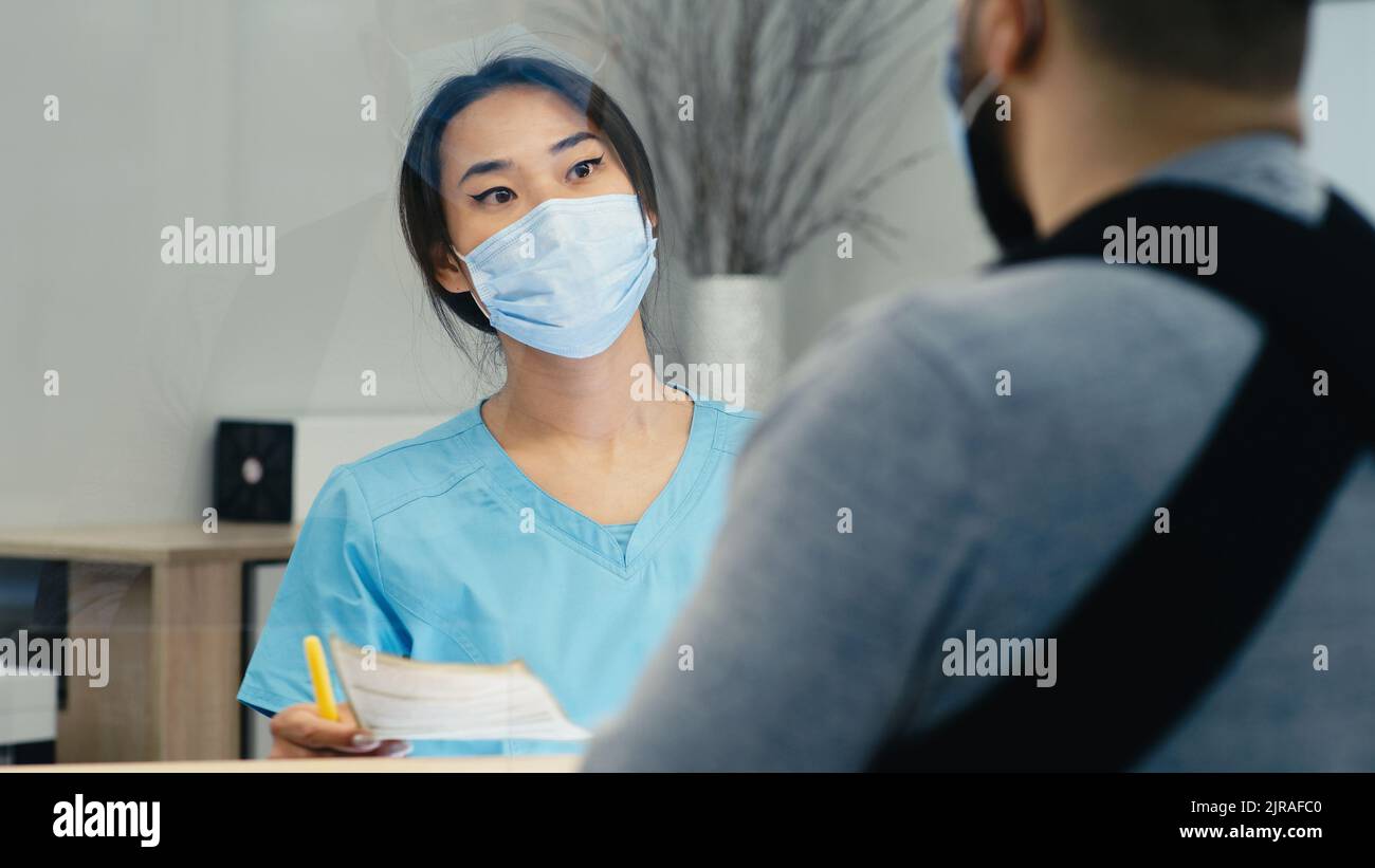 Asian woman in mask giving paper to male patient with broken arm and showing direction while working on reception in modern clinic Stock Photo