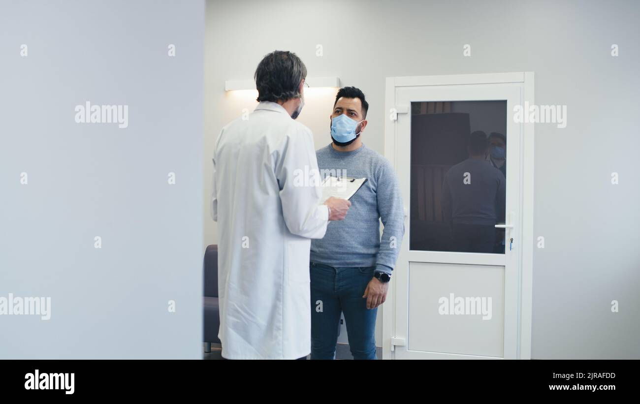Mature medical practitioner in mask reading data from clipboard and talking with man while walking in corridor of contemporary clinic Stock Photo
