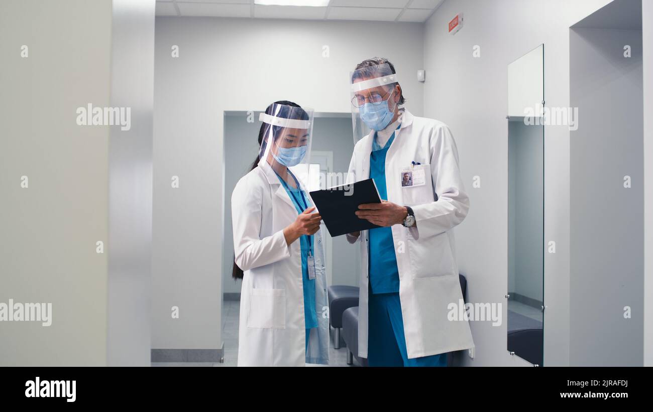 Diverse man and woman in medical uniform and face shields discussing data on clipboard then walking different direction in hallway of contemporary hospital Stock Photo