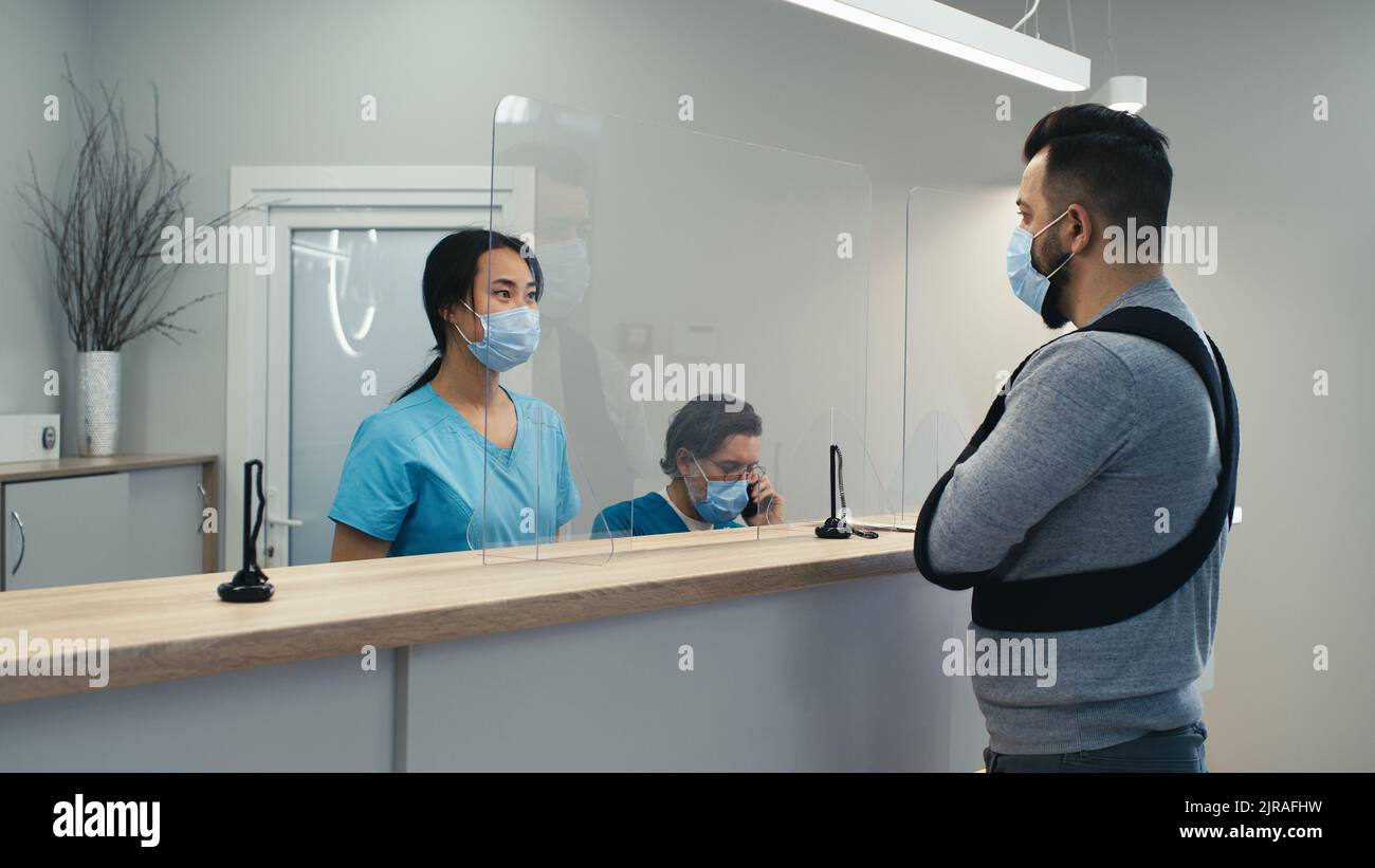 Asian woman giving directions to man with broken arm while working on reception of modern clinic near doctor Stock Photo