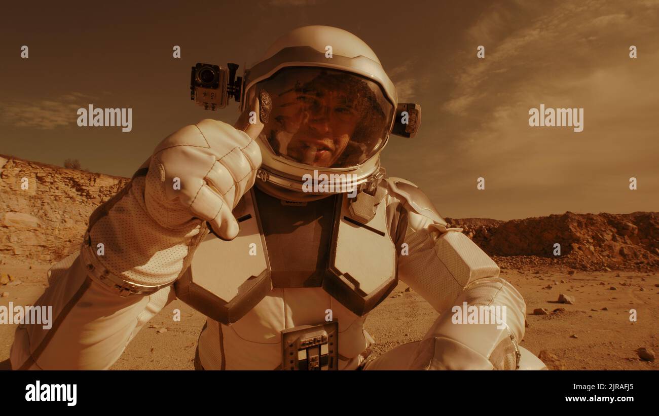 Man in spacesuit walking and turning on camera then waving hand and pointing away while speaking and shooting video during colonization of Mars Stock Photo