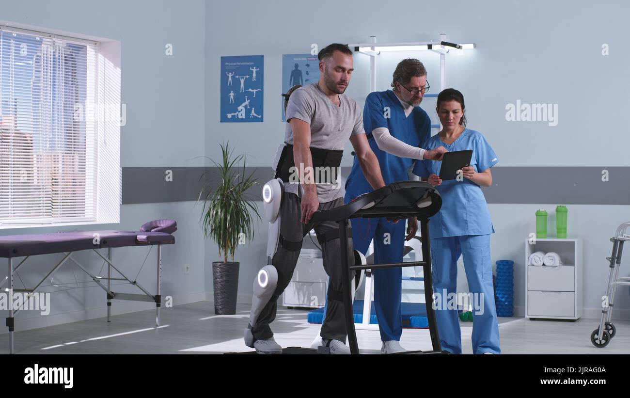 Male and female rehabilitation therapists talking and analyzing data on tablet near man in exoskeleton walking on treadmill in futuristic clinic Stock Photo