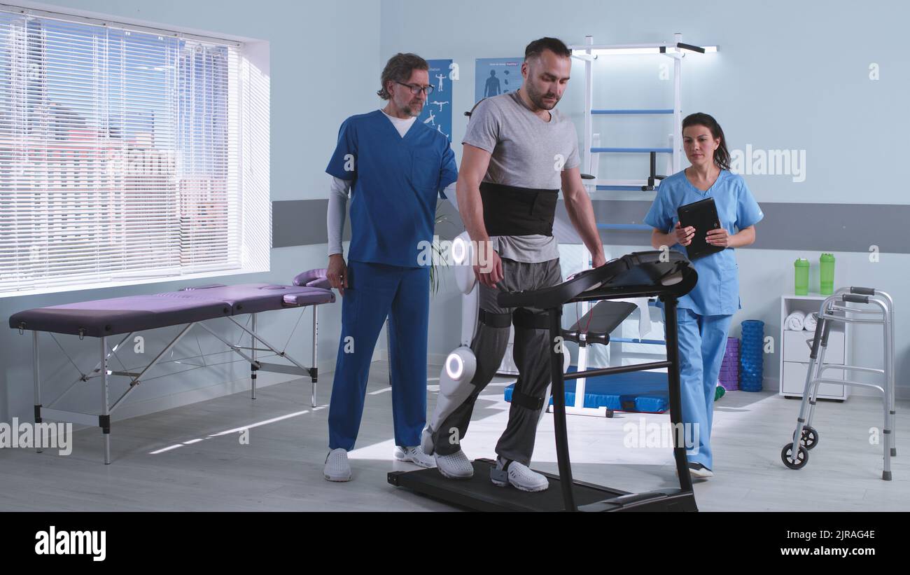 Male and female rehabilitation therapists talking and analyzing data on tablet near man in exoskeleton walking on treadmill in futuristic clinic Stock Photo