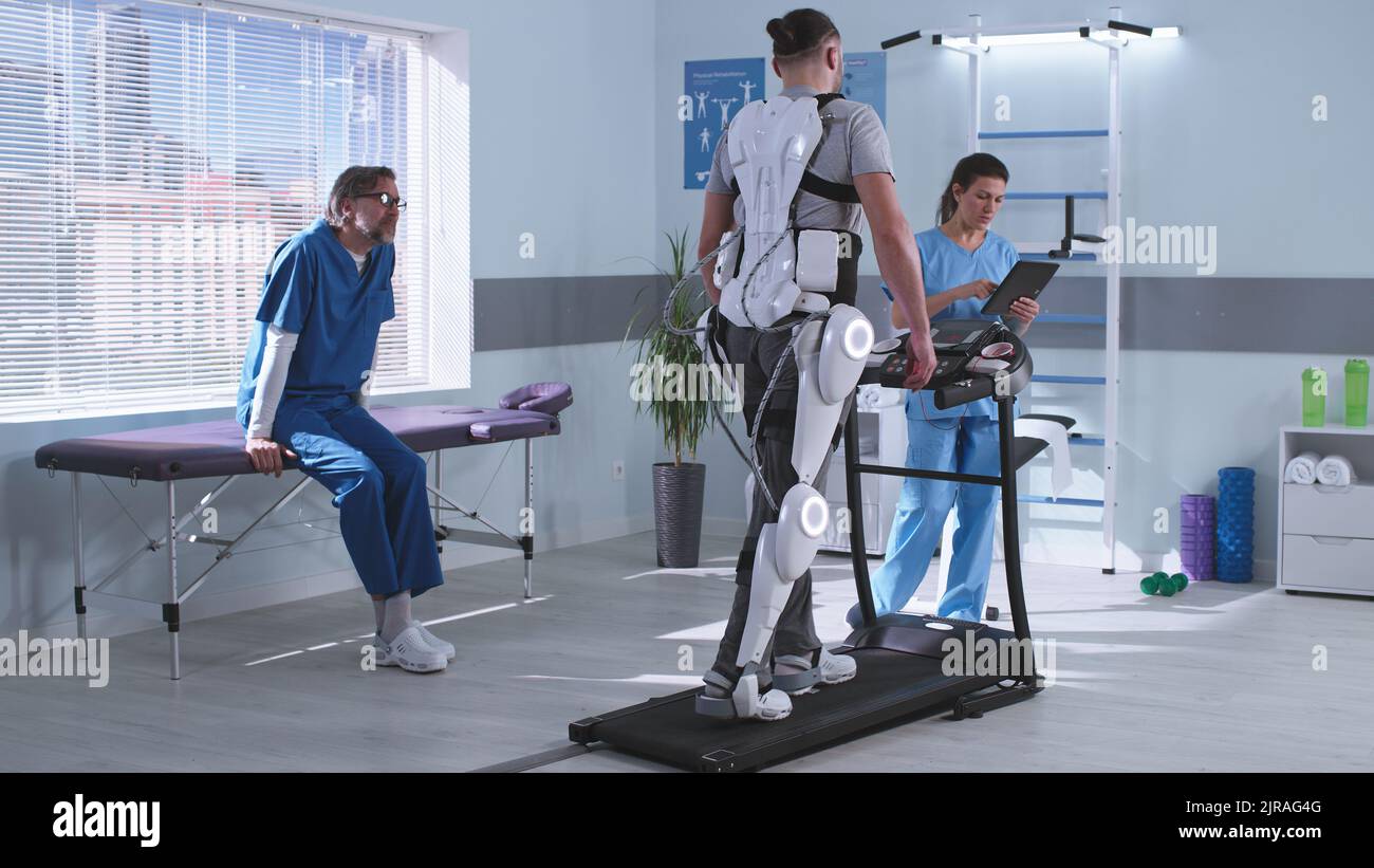 Man and woman in medical uniform controlling treadmill and discussing results while male patient in exoskeleton learning to walk during rehabilitation session in futuristic hospital Stock Photo