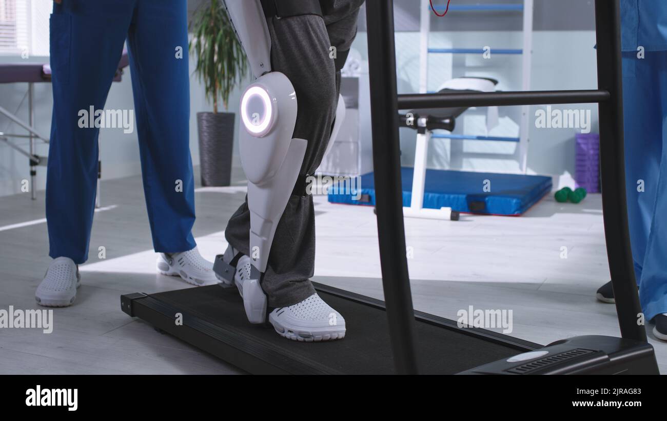 Male patient in exoskeleton walking on treadmill near medical practitioner during rehabilitation session in futuristic hospital Stock Photo