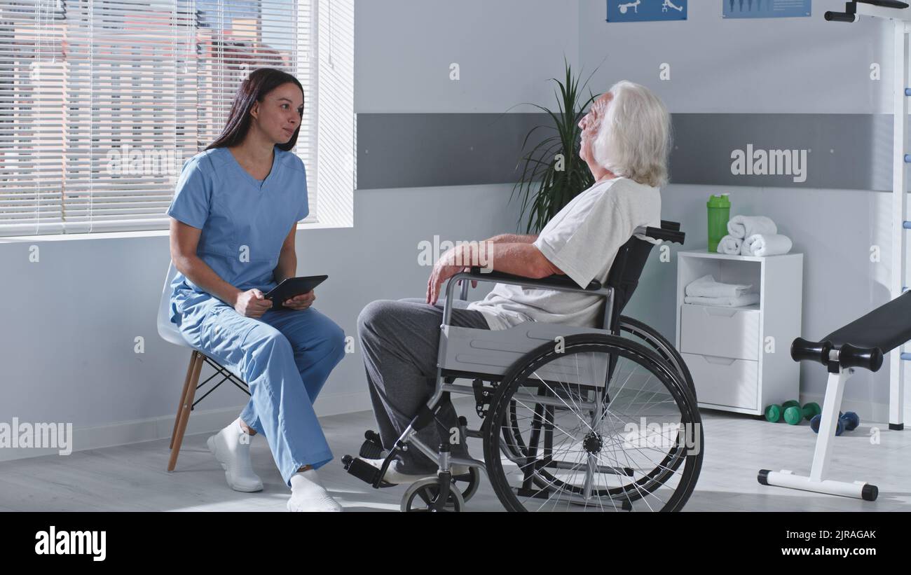 Senior man with disability sitting on wheelchair and speaking with female rehabilitation