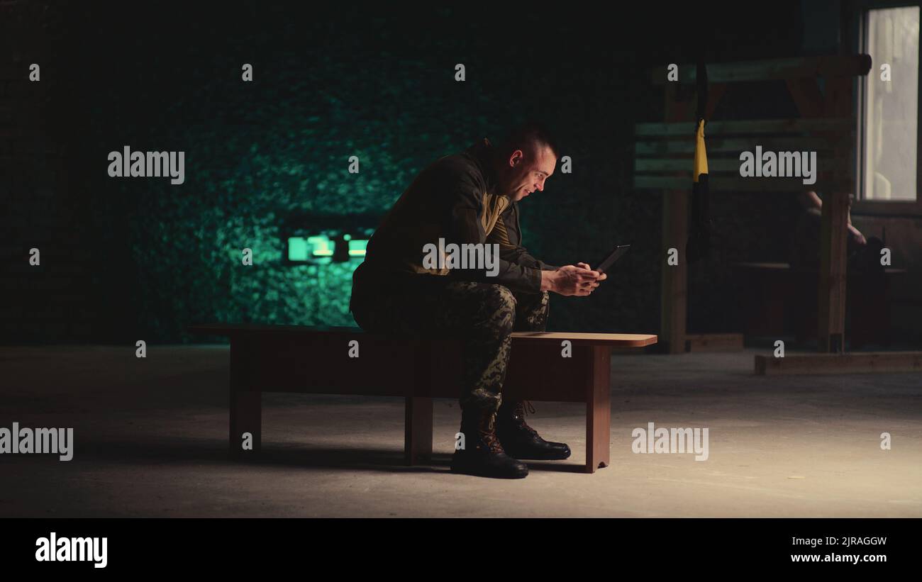 View of military man sitting on bench and making video call on tablet in dark gym Stock Photo