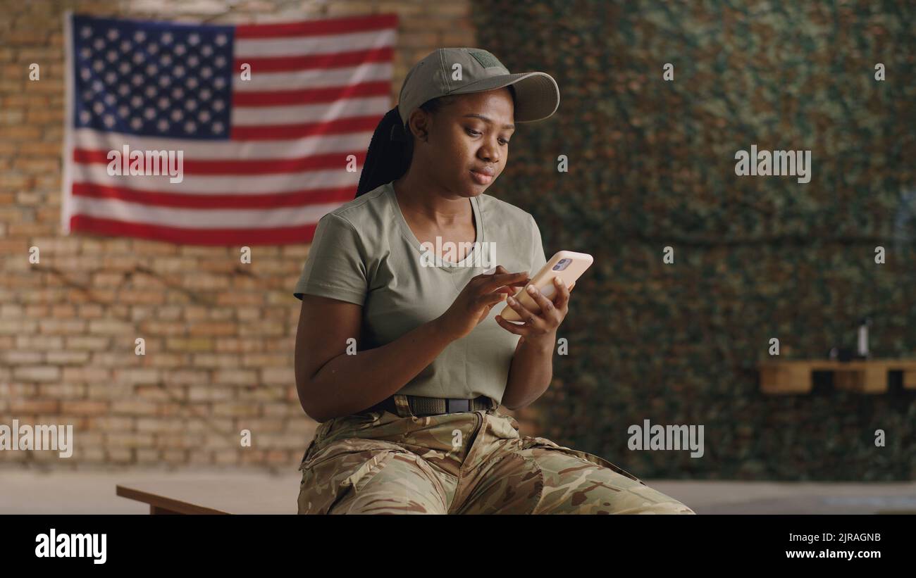 Cheerful black female soldier waving hand and speaking while sitting on bench and making video call to friend in gym of USA military base Stock Photo