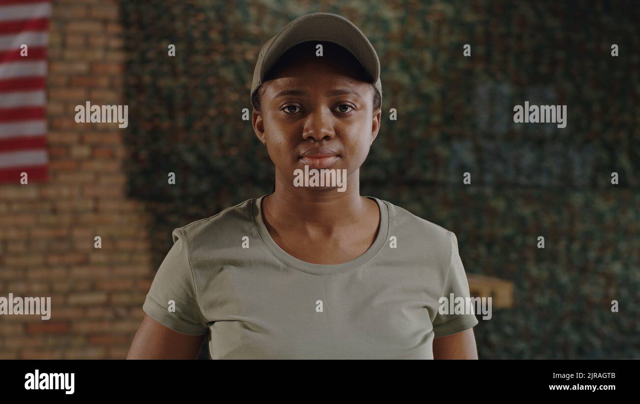 Black female soldier in t shirt and cap looking at camera with serious face while standing against brick wall on USA military base Stock Photo
