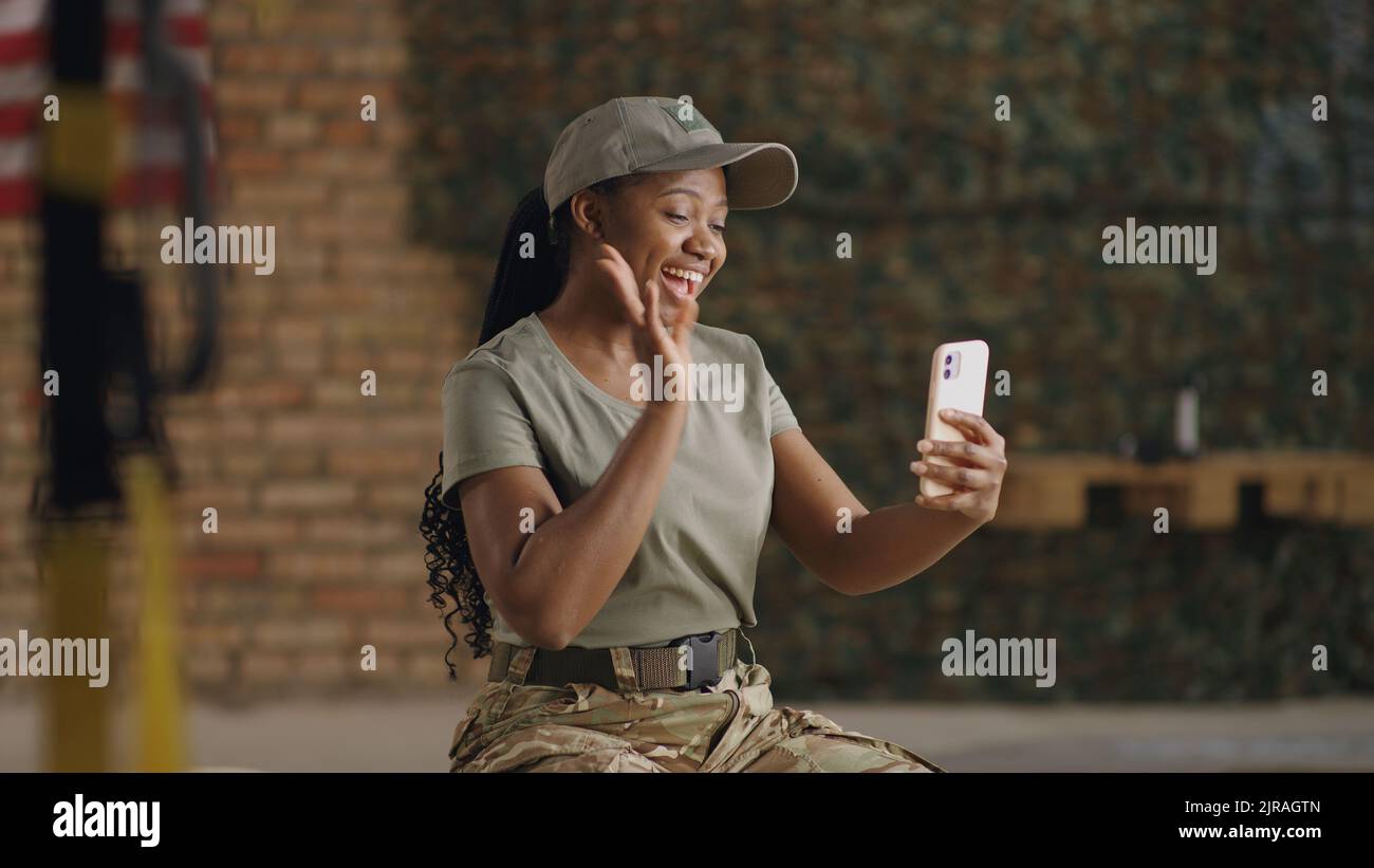 Happy African American female soldier waving hand and smiling while making video call to friend or family on smartphone Stock Photo