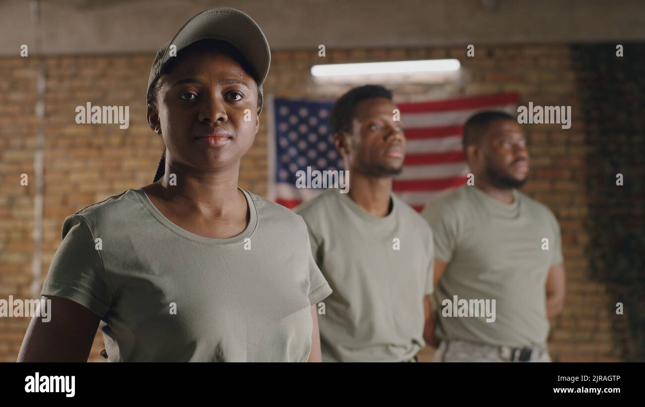 African American female soldier turning head and looking at camera while standing near male squadmates against brick wall with USA flag on military base Stock Photo