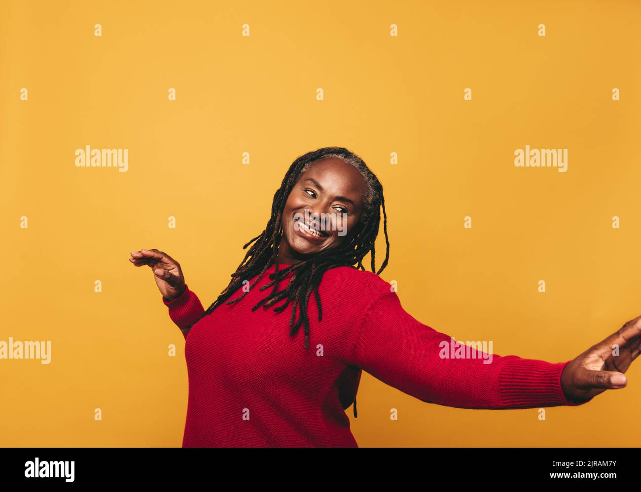 Cheerful mature woman dancing and having fun while standing against a yellow background. Happy black woman with dreadlocks embracing her natural hair Stock Photo