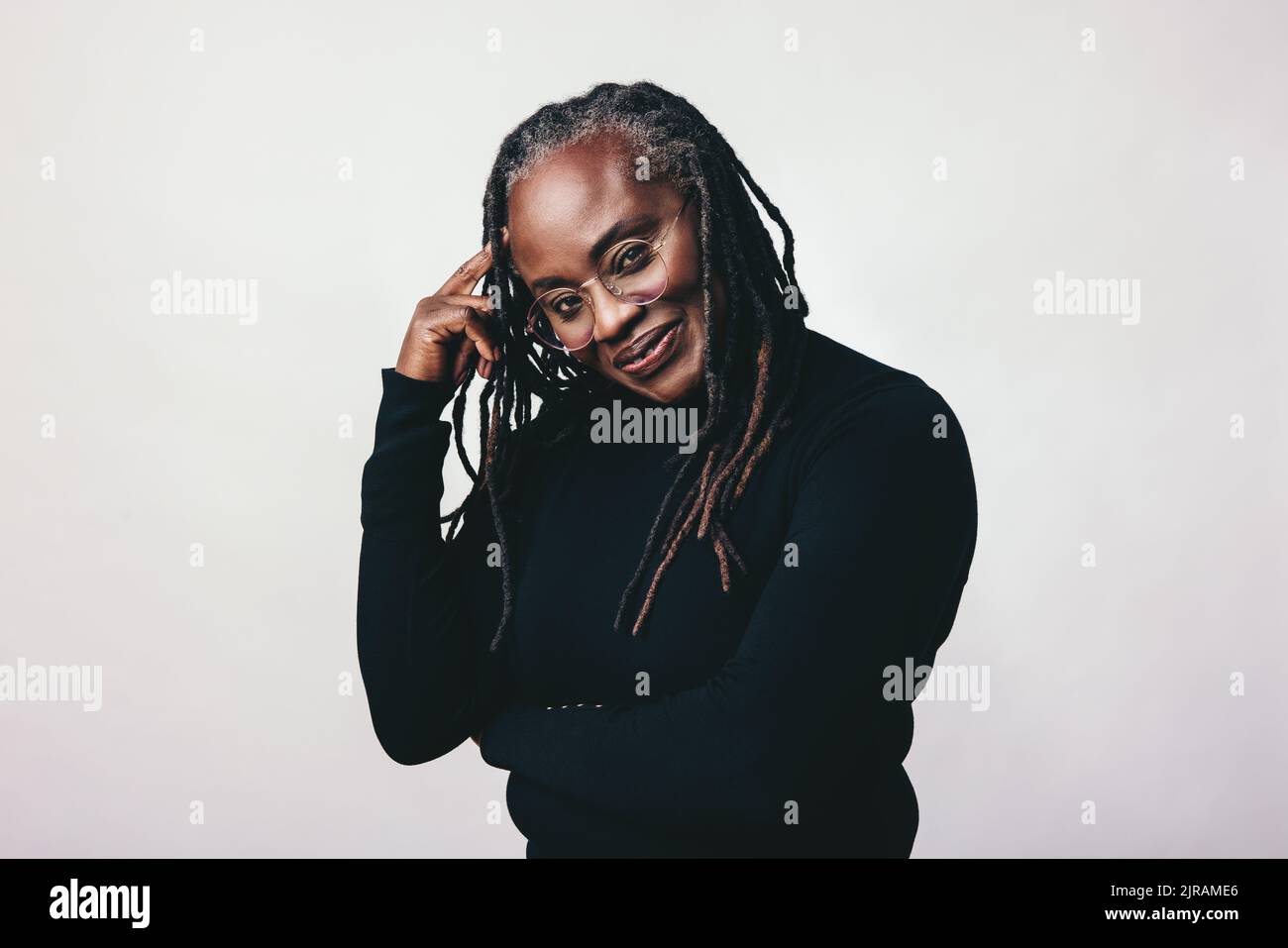 Middle-aged woman looking at the camera while wearing eyeglasses in a studio. Professional woman with dreadlocks standing against a grey background. Stock Photo