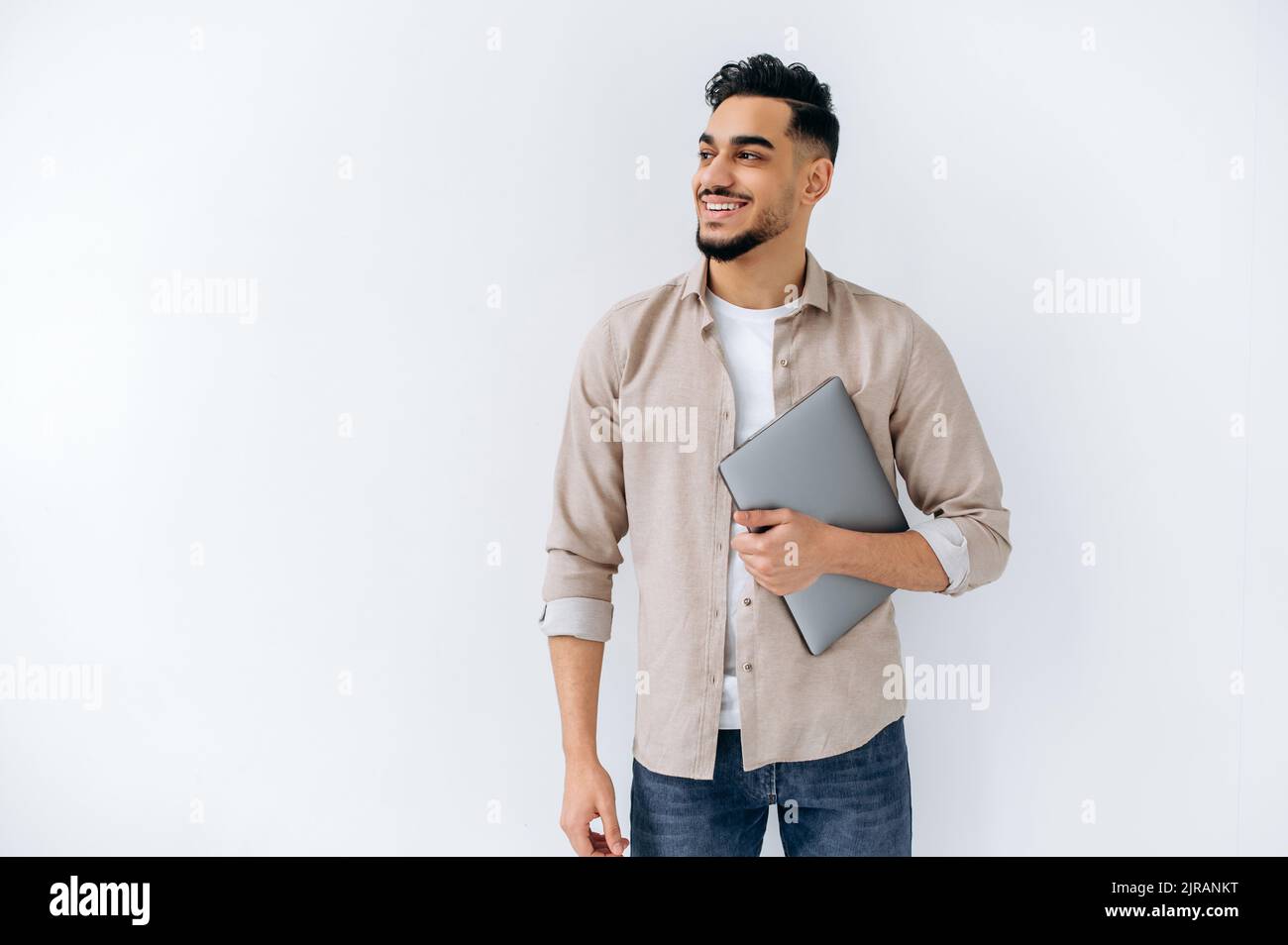 Positive friendly modern arabian or indian young man with beard, wearing casual shirt, standing on isolated white background, holding laptop, looking to the side, smiling, dreaming, thinking Stock Photo