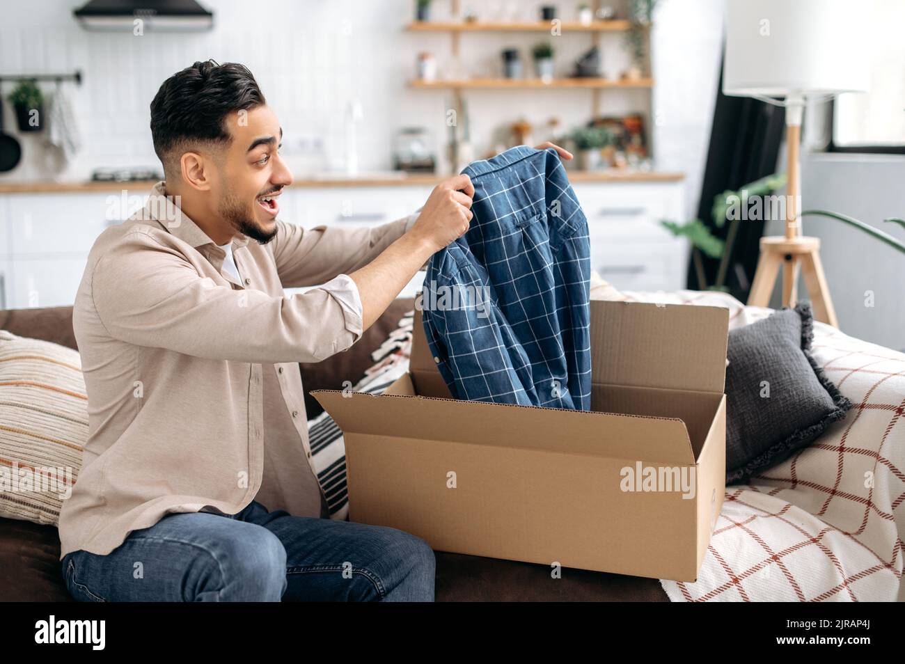 Excited happy indian or arabian guy opening big carton box, unpacking internet store order at home, contented with fast delivery service and the goods which received, smiling and feel satisfied Stock Photo