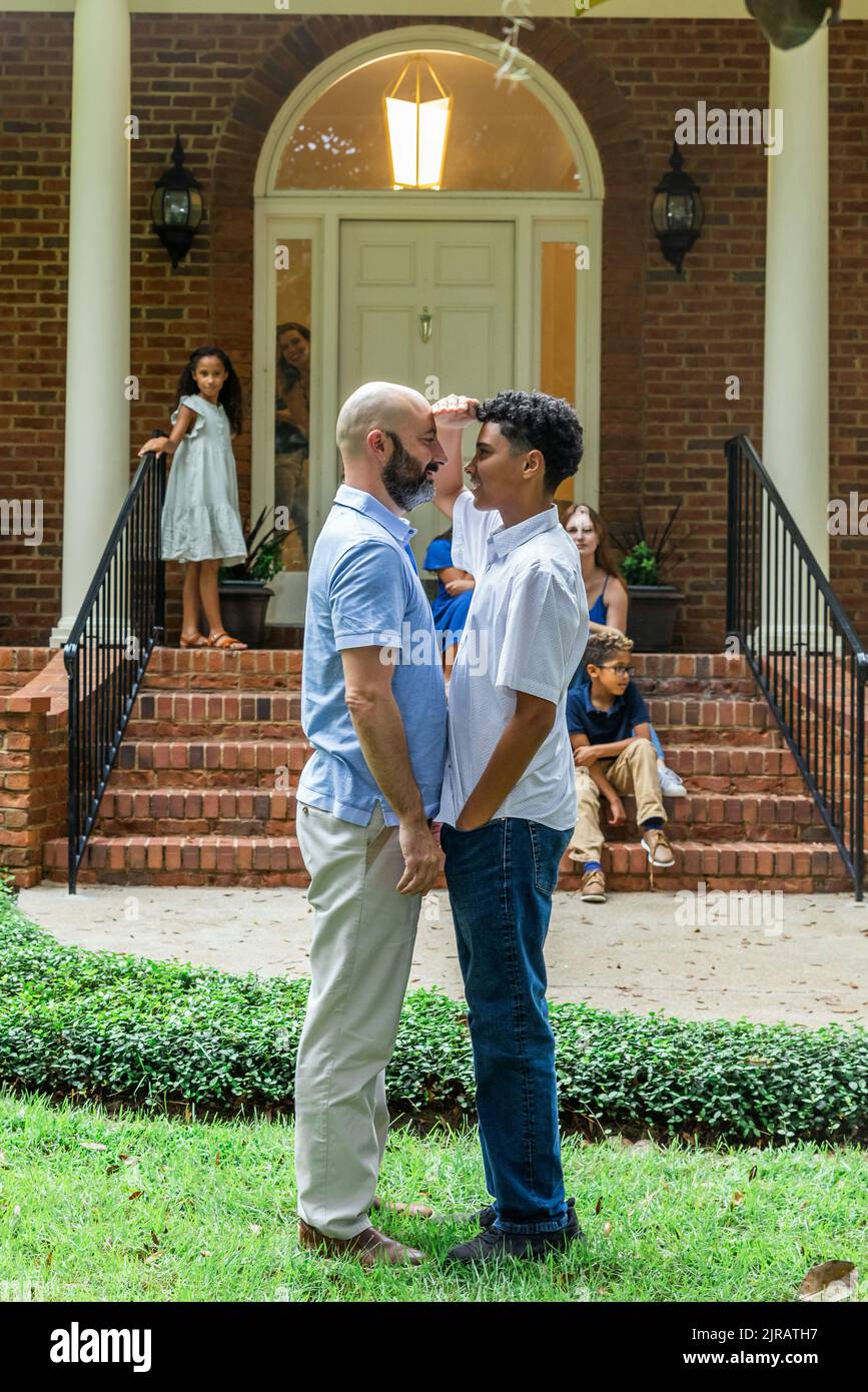 A teenage son showing his father that he is almost as tall as he is by measuring their height outside their family home with the family looking on Stock Photo