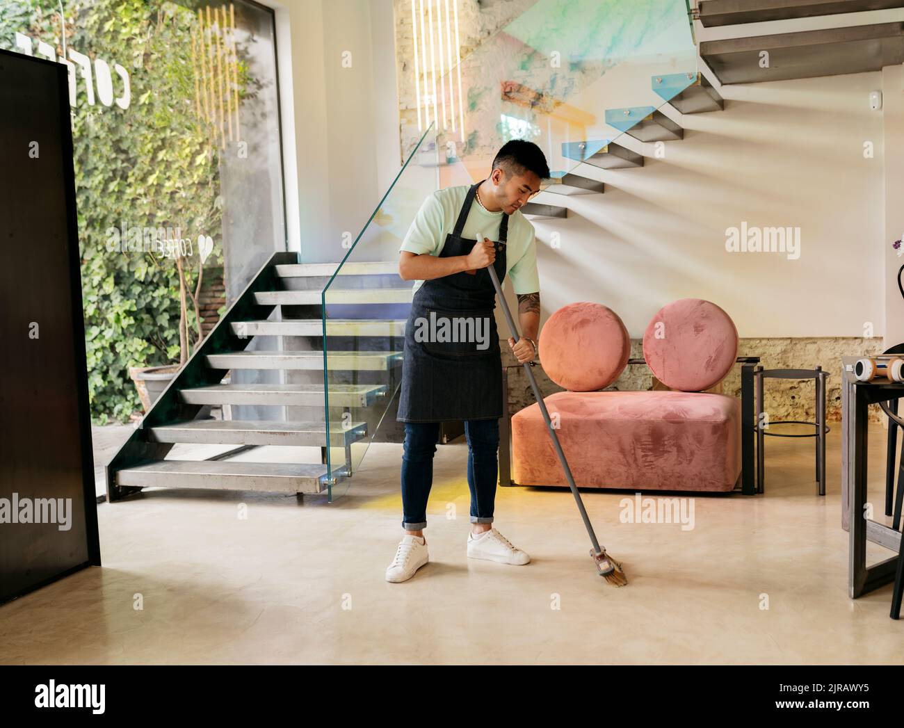 Young man wearing apron cleaning floor with broom in cafe Stock Photo