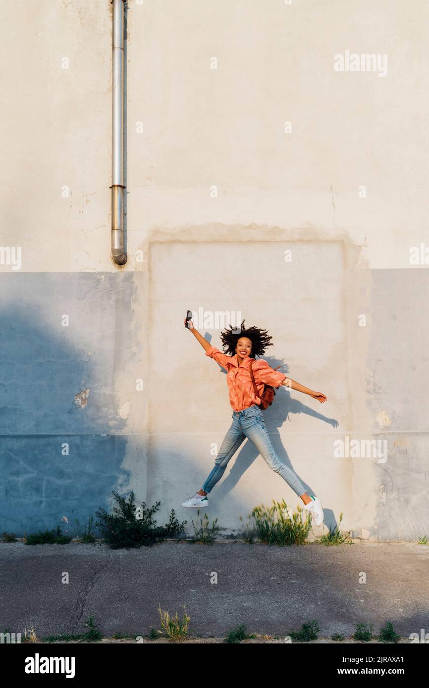 Happy woman holding smart phone jumping in front of wall Stock Photo ...