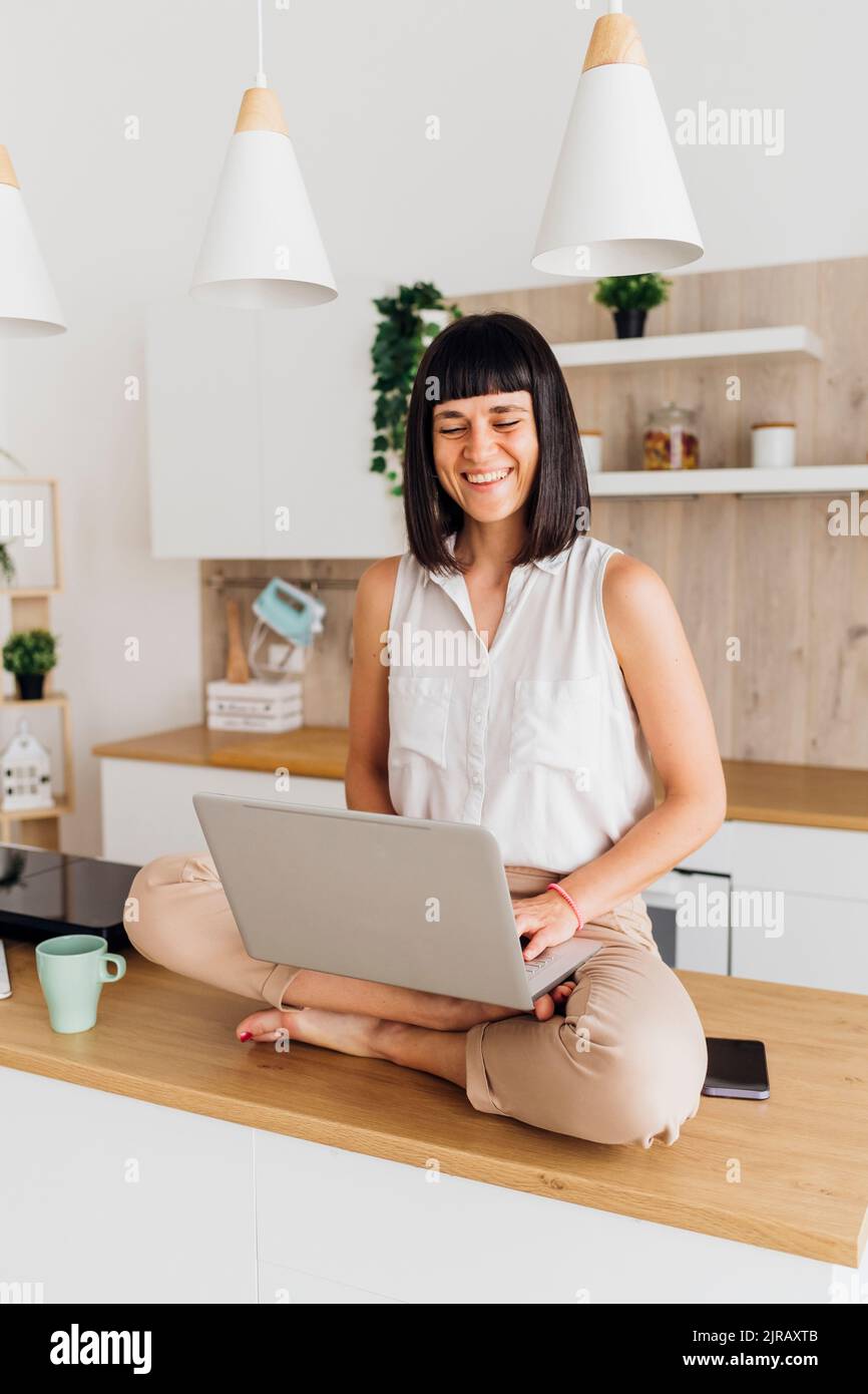 Smiling freelancer with laptop sitting on kitchen island at home Stock Photo