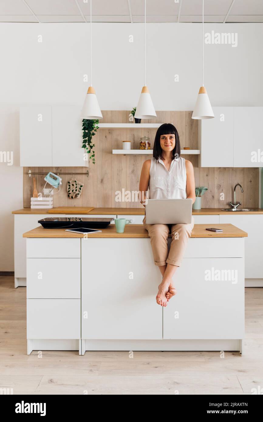 Woman with laptop sitting on kitchen island at home Stock Photo