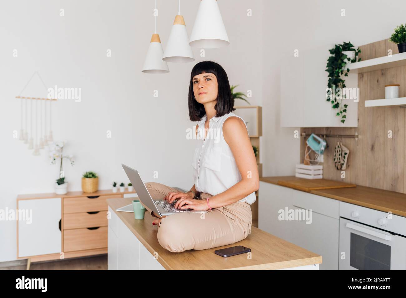 Thoughtful freelancer sitting on kitchen island with laptop at home Stock Photo